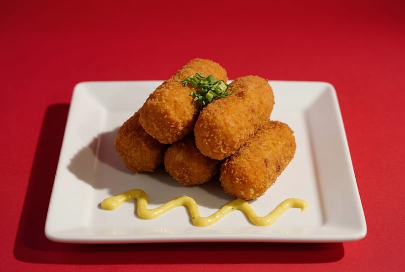 Golden-brown croquettes topped with chives and a squiggle of mustard on a white rectangular plate.