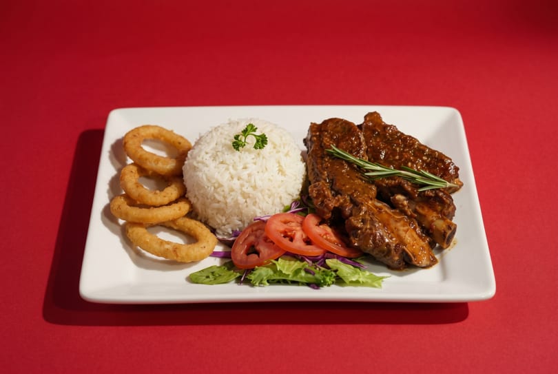 Plate of BBQ ribs with rice, onion rings, and a side salad on a red background.