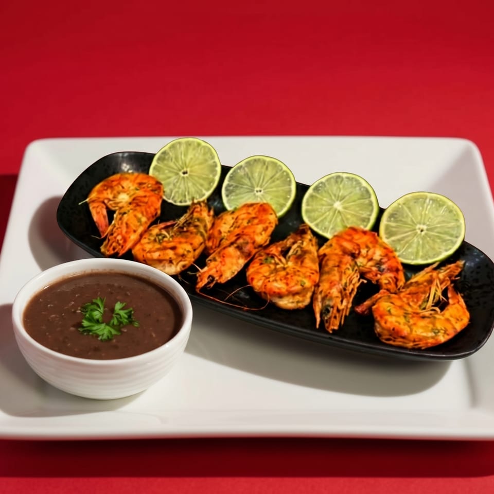 Grilled shrimp with lime slices on a black plate, next to a bowl of dark sauce, on a white platter.