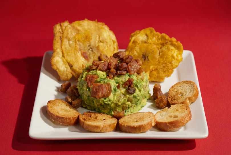 Guacamole topped with bacon bits, served with plantain chips and toasted bread slices on a white plate.