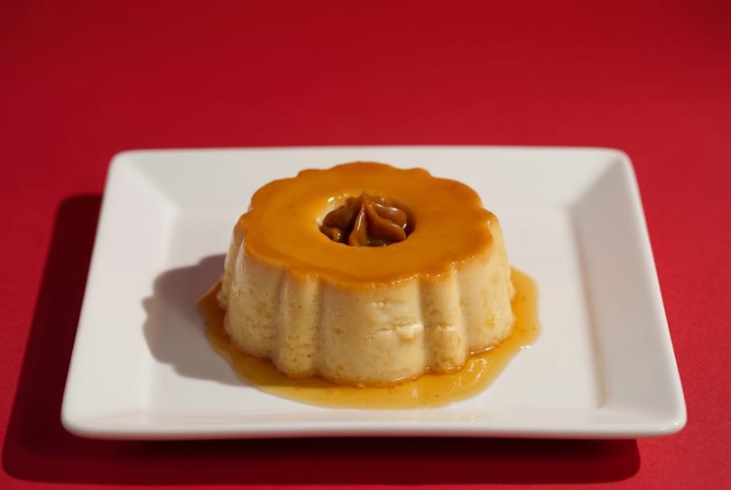 Flan dessert with dulce de leche in the center, on a white plate against a red background.